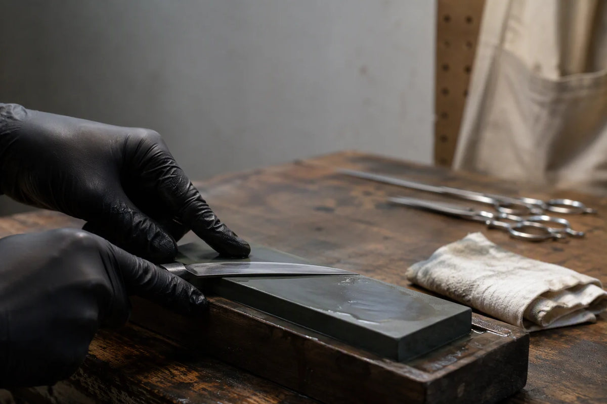 Gloved hands honing a grooming shear blade flat on a wet whetstone at a workshop bench